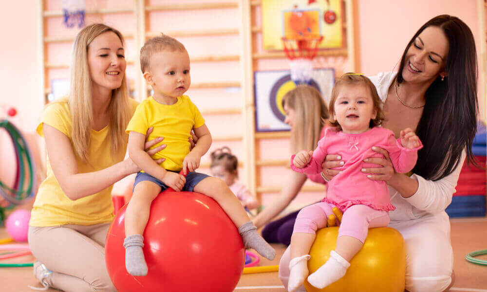 Two year olds sitting on gym balls assited by instructors